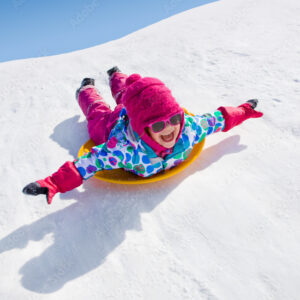 Enfant souriant avec bonnet rose et lunettes de soleil descendant une piste enneigee en luge lors d’une colonie de vacances d’hiver à Neige et Soleil
