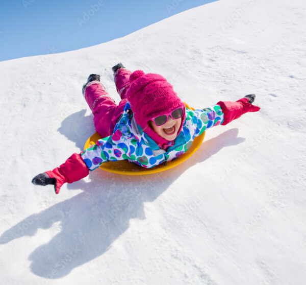 Enfant souriant avec bonnet rose et lunettes de soleil descendant une piste enneigee en luge lors d’une colonie de vacances d’hiver à Neige et Soleil